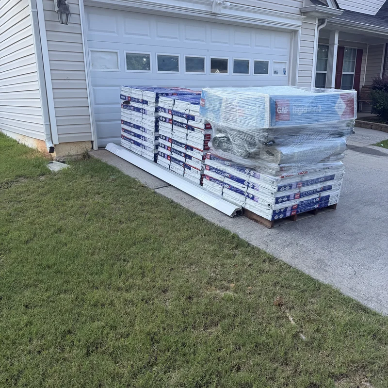 Wrapped pallets of GAF Timberline shingles and roofing accessories delivered beside a garage.