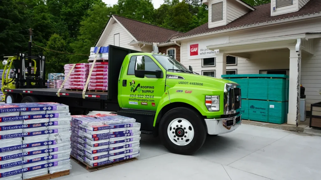 A green A+ Roofing Supply flatbed truck beside pallets of shingles.