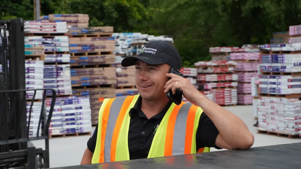 A person wearing a reflective vest speaks on a phone in front of stacked shingles.